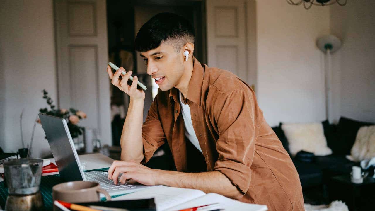 Young male freelancer talking on speaker phone while working on laptop at home office