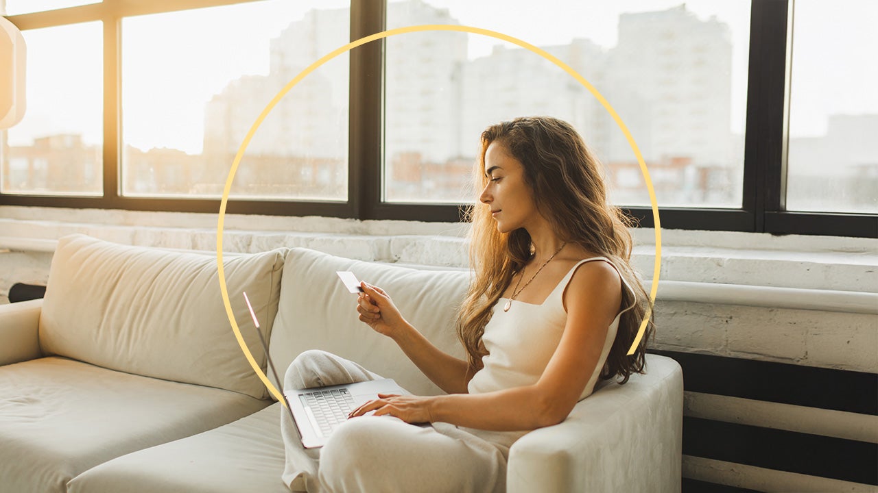 A young woman sits on a white couch looking at her credit card with a laptop on her lap. There is an illustrated yellow circle behind her.