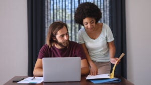 A Black woman and a white man work together. The woman is standing and reviewing a book while the man is on a laptop.