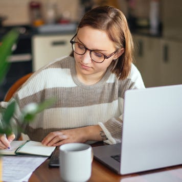 Young white woman working at a laptop and writing in a notebook at her kitchen table.