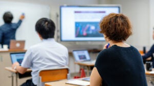 Two adult learners shown from behind along with a teacher at the whiteboard.