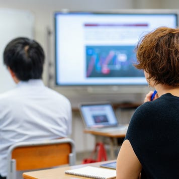 Two adult learners shown from behind along with a teacher at the whiteboard.