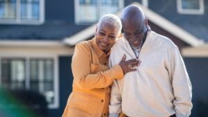 A wealthy retired couple embraces outside their home.