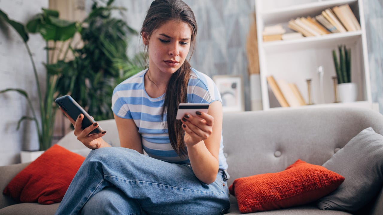 A young woman sits on a couch with a focused expression, holding a phone and a credit card in her hands, engaging in an online transaction.