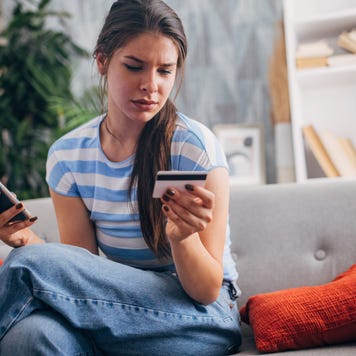 A young woman sits on a couch with a focused expression, holding a phone and a credit card in her hands, engaging in an online transaction.