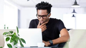 Young Black man in glasses sitting at a table and looking thoughtful while using his laptop
