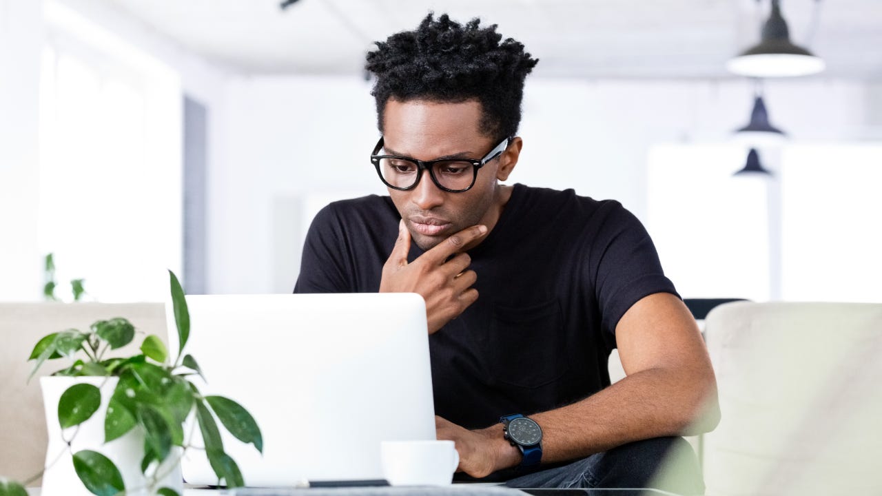Young Black man in glasses sitting at a table and looking thoughtful while using his laptop