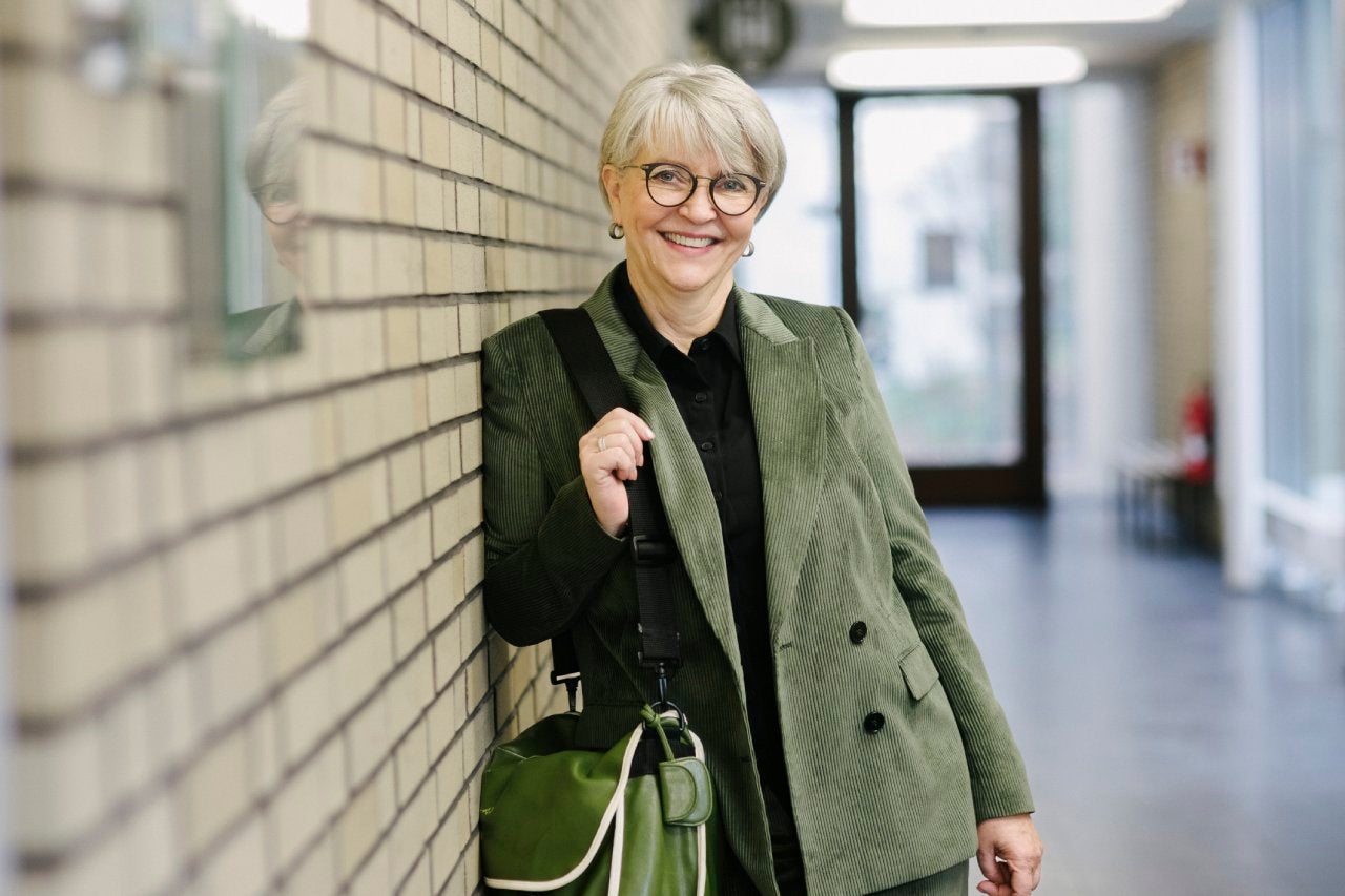 A portrait of a mature school tutor smiling while leaning on a brick wall in a school hallway.