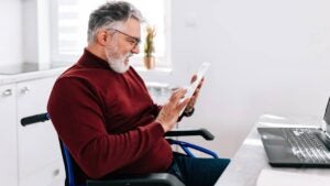 A mature Caucasian man in a wheelchair is cheerfully using his tablet, while sitting at his home desk.