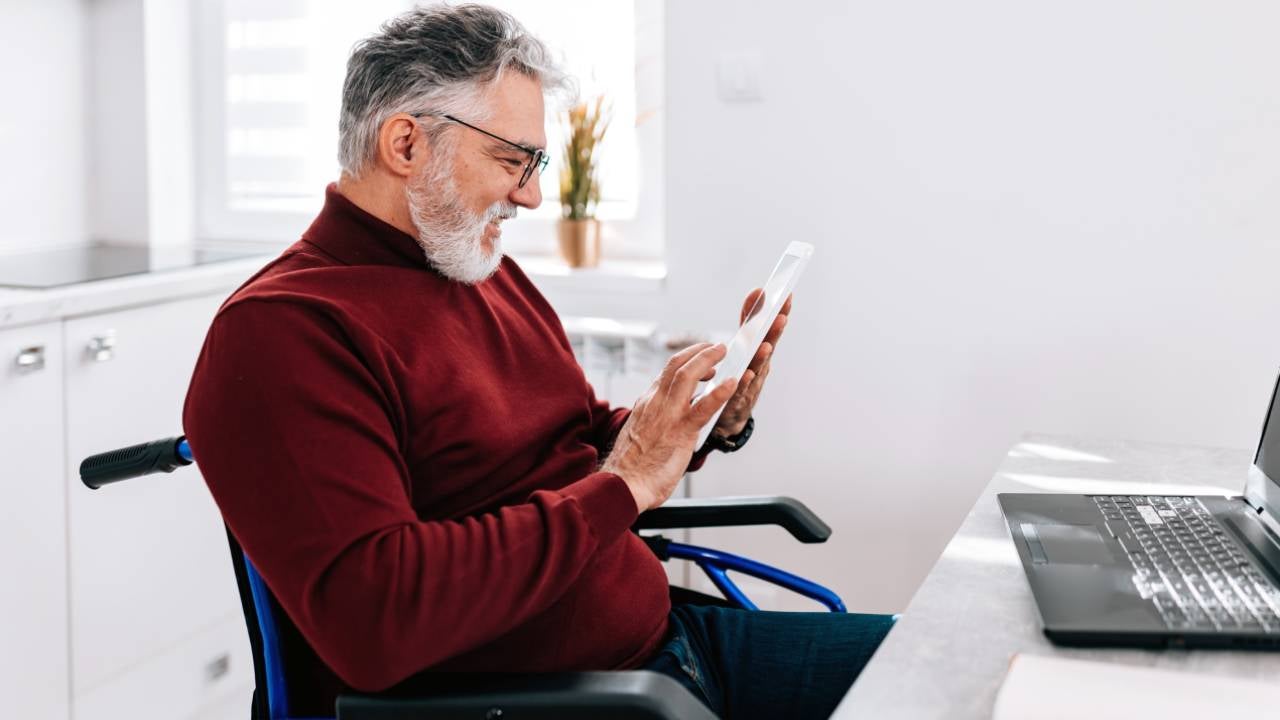 A mature Caucasian man in a wheelchair is cheerfully using his tablet, while sitting at his home desk.
