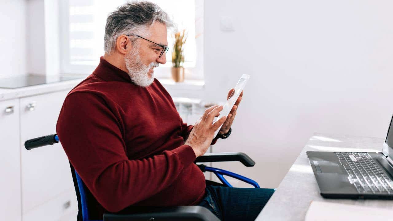 A mature Caucasian man in a wheelchair is cheerfully using his tablet, while sitting at his home desk.