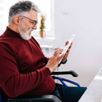 A mature Caucasian man in a wheelchair is cheerfully using his tablet, while sitting at his home desk.