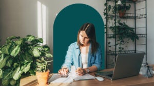 A white woman sits in a home office surrounded by plants while she does paperwork.