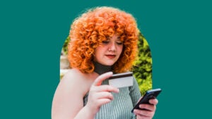 A woman with red curly hair looking down at a credit card in her right hand with a cellphone in her left hand.