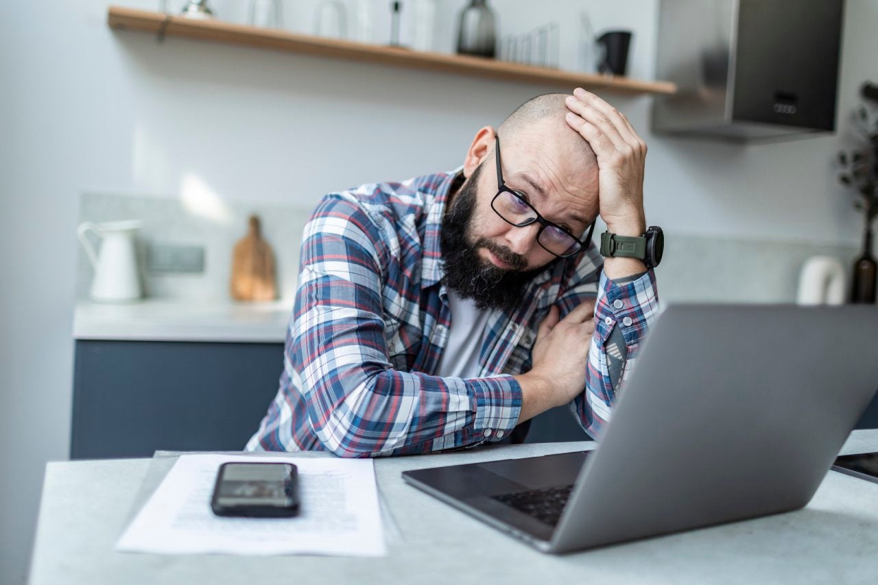 Adult man sitting at the kitchen with laptop and looking upset with the news.