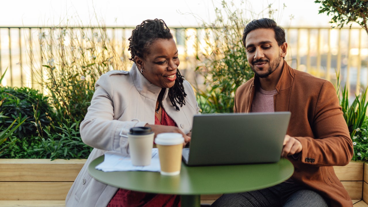 A woman and a man sit outside at a small table with a laptop and coffee in front of them. Both are smiling as they talk.
