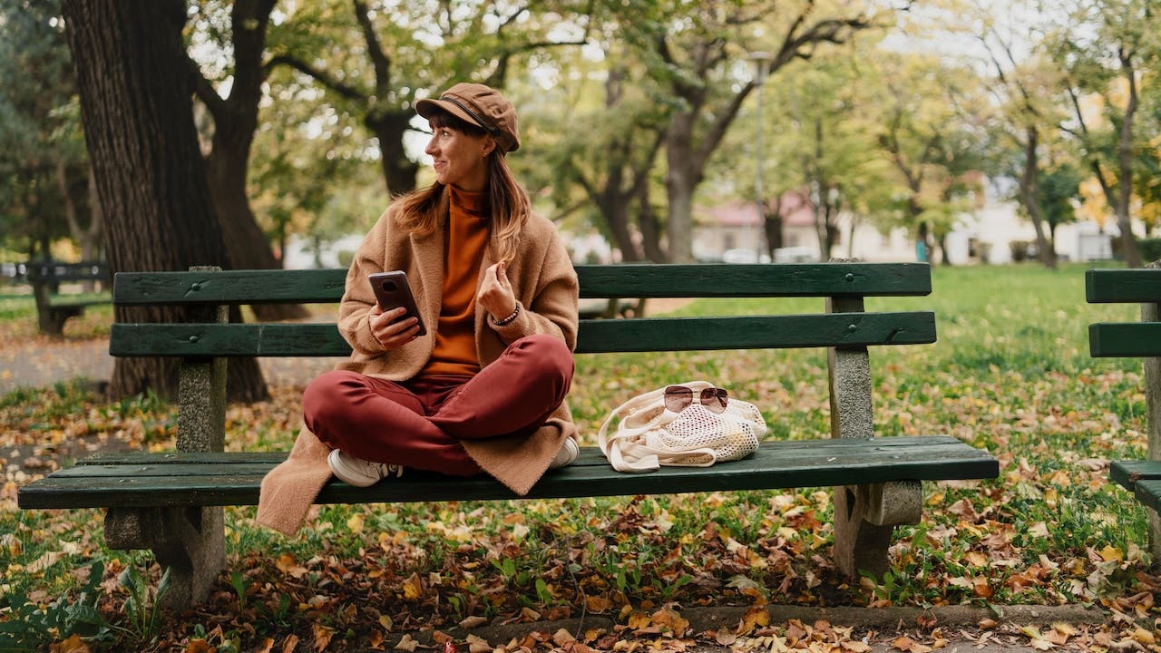 Mid-adult woman sitting on bench and using mobile phone outside in the city on autumn day.