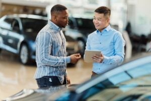 Smiling Asian salesman talking to a Black customer while using digital tablet in a car showroom.