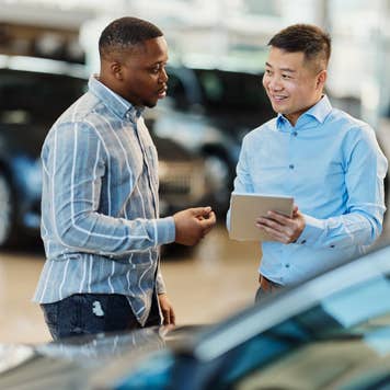 Smiling Asian salesman talking to a Black customer while using digital tablet in a car showroom.