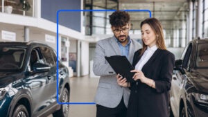 A man and a woman in suits stand together in a dealership while looking at a tablet.