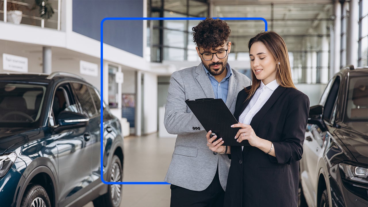 A man and a woman in suits stand together in a dealership while looking at a tablet.
