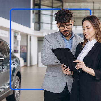 A man and a woman in suits stand together in a dealership while looking at a tablet.