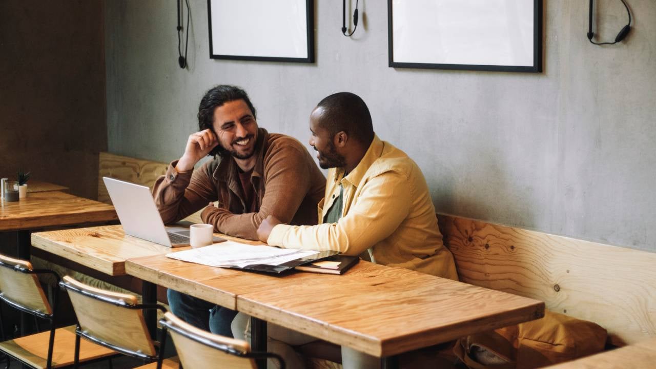 Two men talk over a laptop and paperwork at a coffee shop.