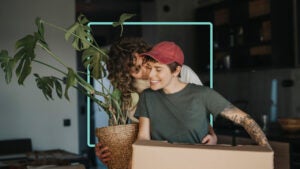 A woman is kissing her partner’s cheek while carrying a plant. Her partner is carrying a brown moving box.