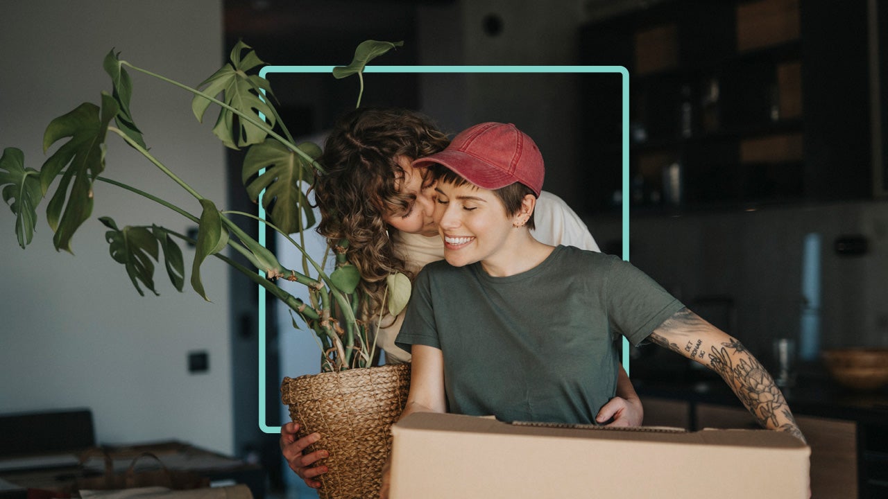 A woman is kissing her partner’s cheek while carrying a plant. Her partner is carrying a brown moving box.