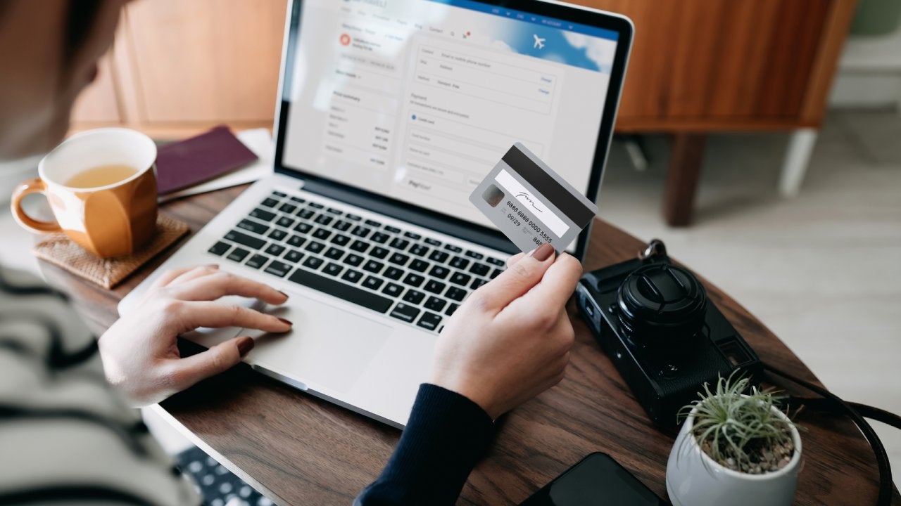 Over the shoulder view of young Asian woman shopping online for flight tickets on airline website with laptop, entering credit card details to make mobile payment at home. Camera and passport on the table. Travel planning. Booking a holiday online