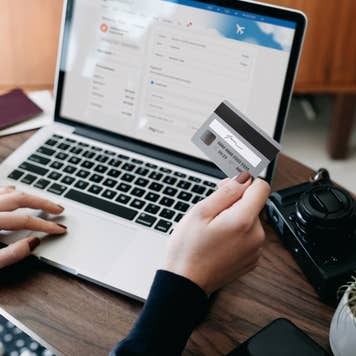 Over the shoulder view of young Asian woman shopping online for flight tickets on airline website with laptop, entering credit card details to make mobile payment at home. Camera and passport on the table. Travel planning. Booking a holiday online