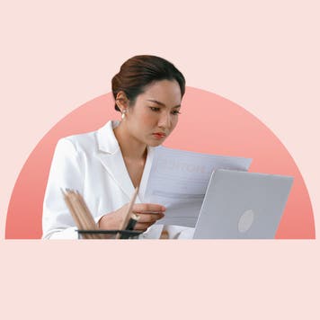 Young woman focused on reading paperwork while looking at laptop screen.