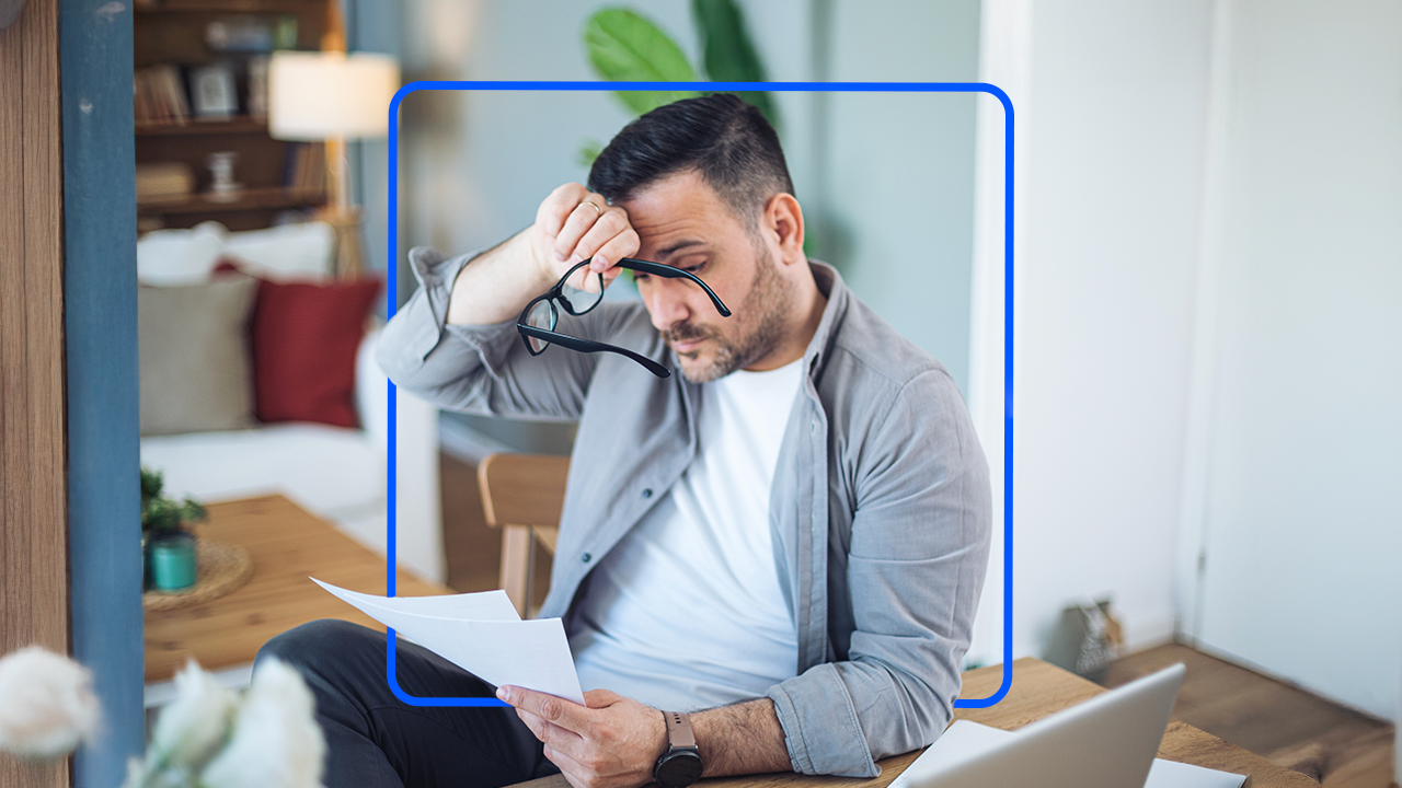 A man looks distressed while reviewing a document. He is holding a pair of black glasses and resting the back of his wrist against his forehead.
