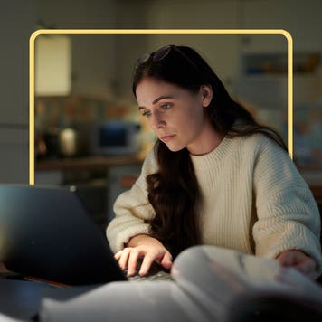 Woman with long dark hair sitting at a table working on a laptop computer that is surrounded by open binders and books.