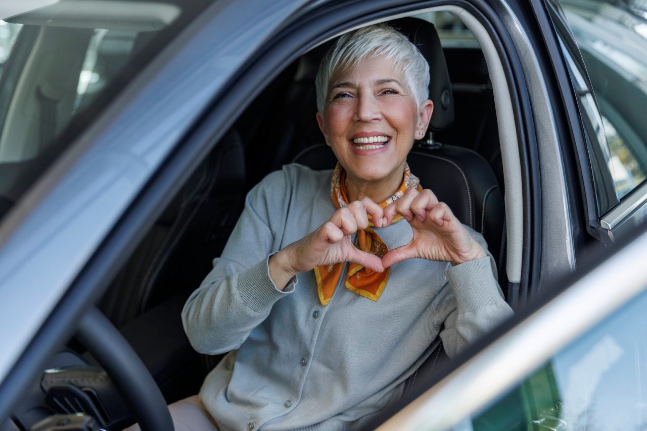 Happy woman gesturing heart shape sitting in car at showroom