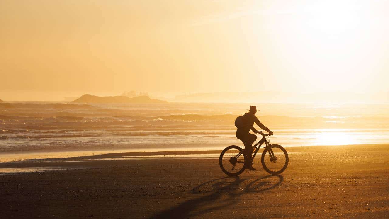 A woman goes for a bike ride at low tide on Long Beach on Vancouver Island near Tofino, British Columbia, Canada, at sunset. She wears casual clothing, a sunhat, and a backpack.