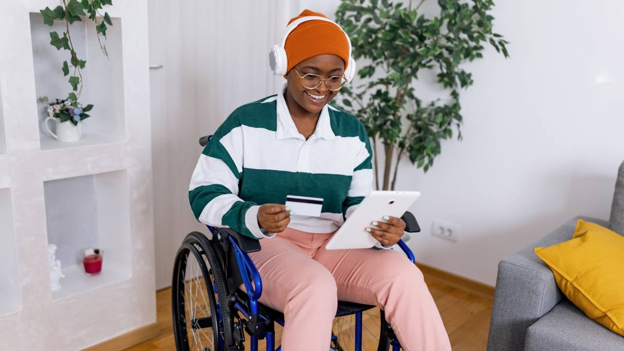 A young African American woman in a wheelchair is holding her tablet and credit card while shopping online.