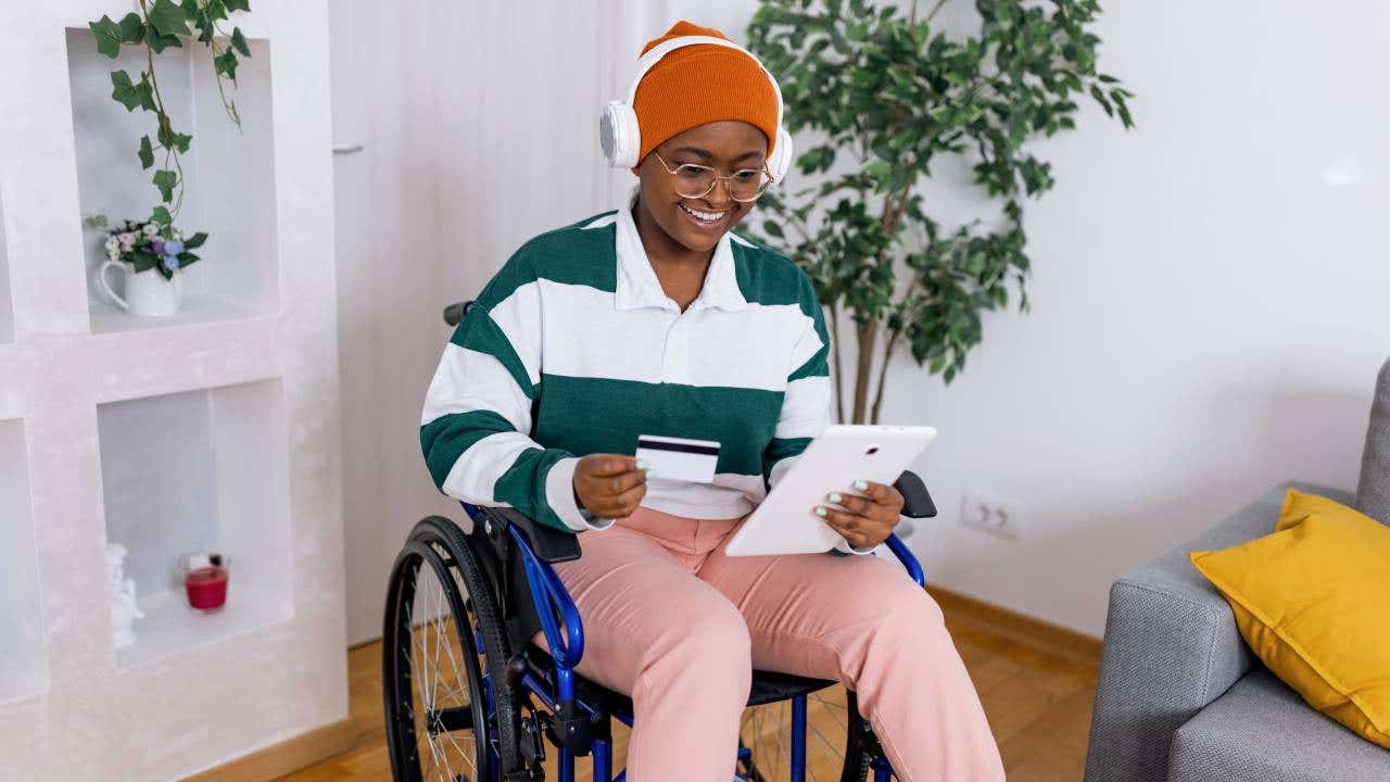 A young African American woman in a wheelchair is holding her tablet and credit card while shopping online.