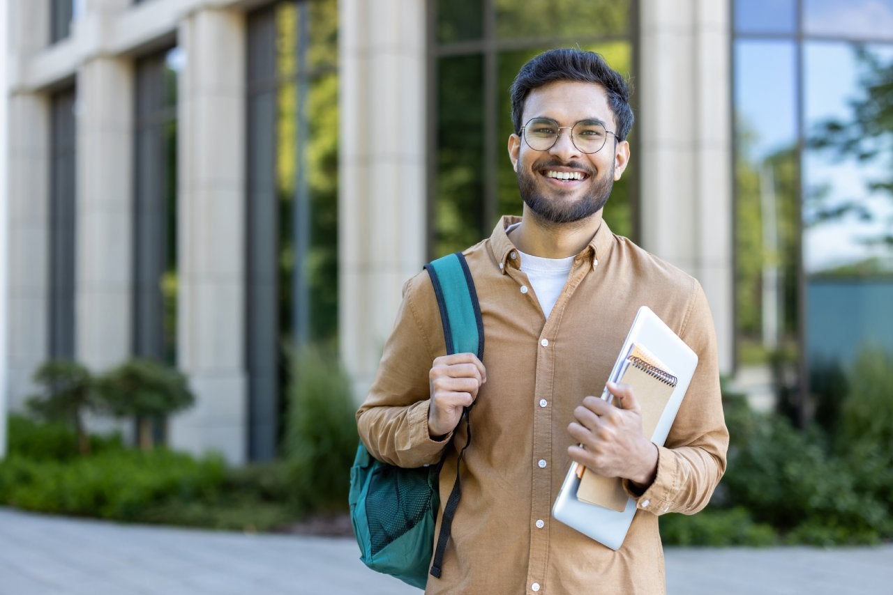 Smiling hispanic student stands outside academic building holding backpack and notebooks. Concept of higher education, confidence, and youth. Ideal for educational and lifestyle themes.