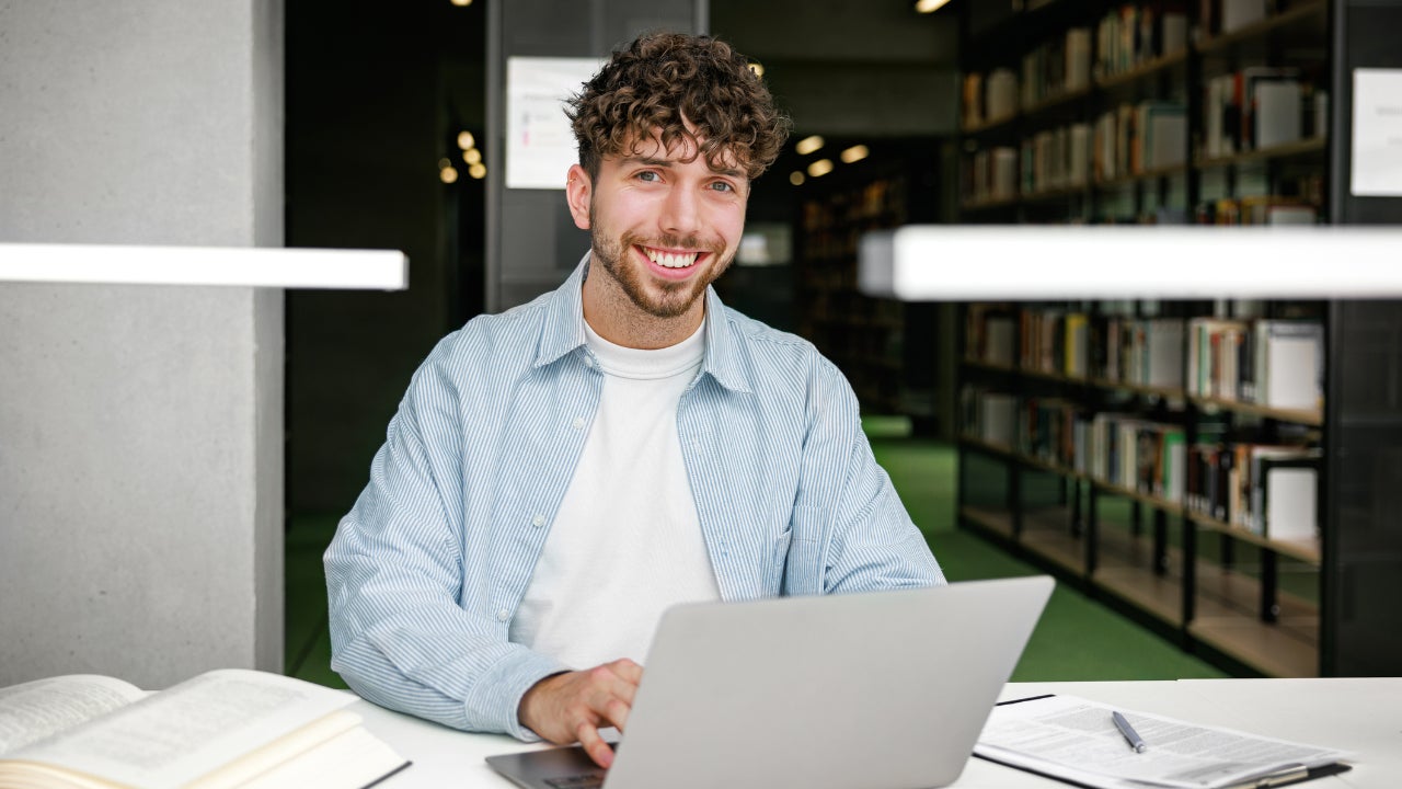 A portrait of a smiling young man works on his laptop with open books and notes nearby in a library. The inviting atmosphere and organized shelves emphasize focus and positivity in learning.