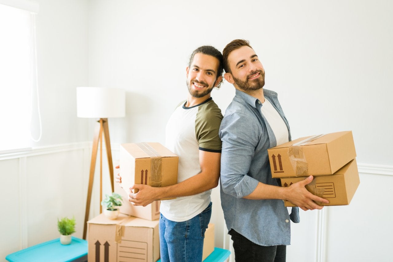 Smiling attractive man and his gay partner looking happy while carrying boxes to move in together