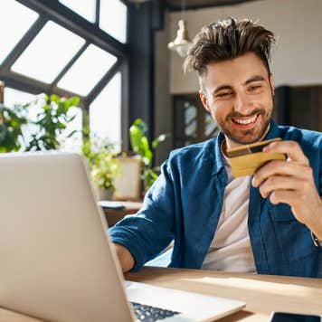 Man sits in front of laptop holding credit card and smiling