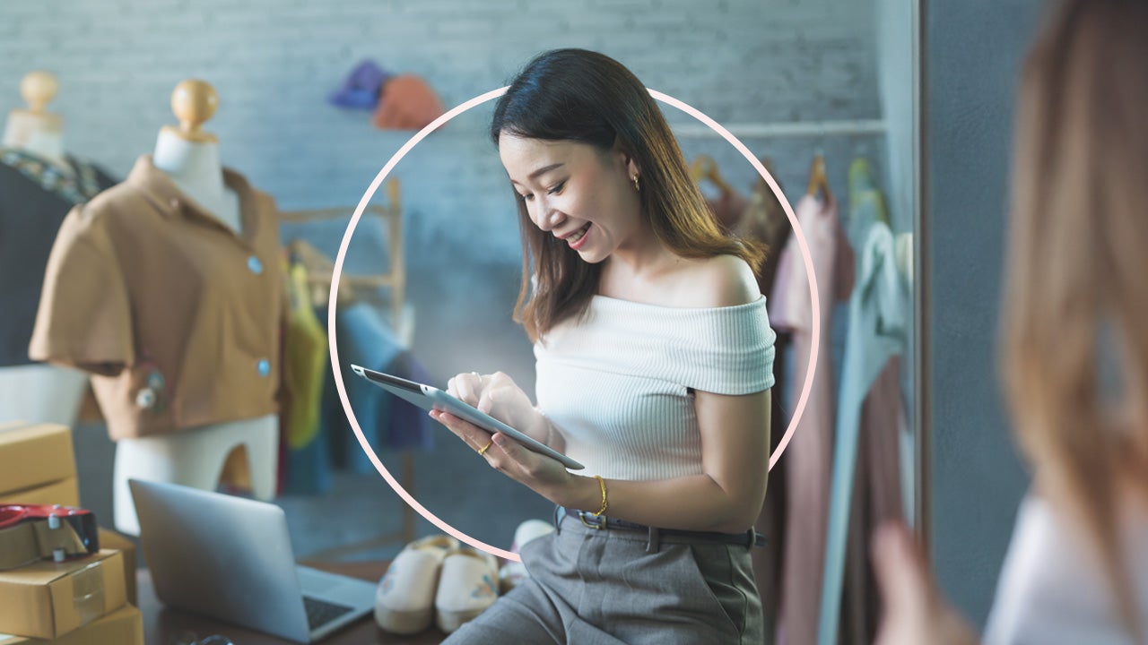 A woman, with shoulder length brown hair, leans on a desk while looking at the tablet in her hands.