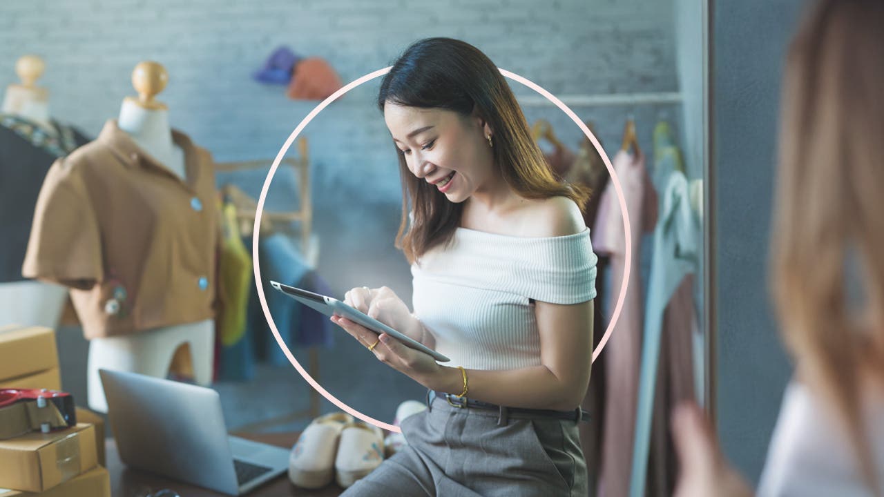 A woman, with shoulder length brown hair, leans on a desk while looking at the tablet in her hands.