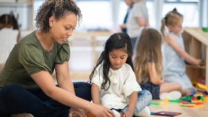 A female preschool teacher of African ethnicity helps one of her students build a tower using magnetic tiles.