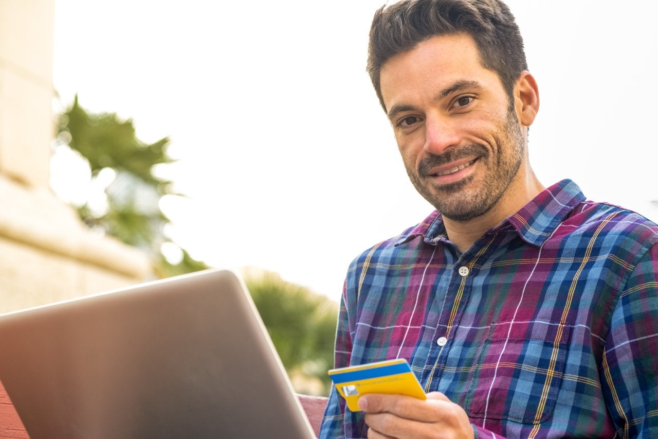 A man makes purchases with a credit card on his laptop in a city park.
