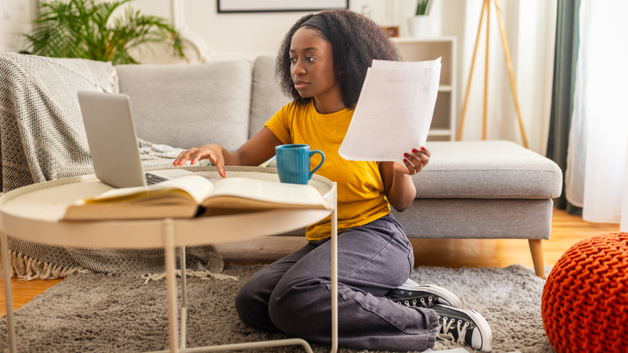 A focused woman in a yellow shirt examines a bill while working on her laptop, sitting on the floor with various documents around her, in a welcoming living room setup