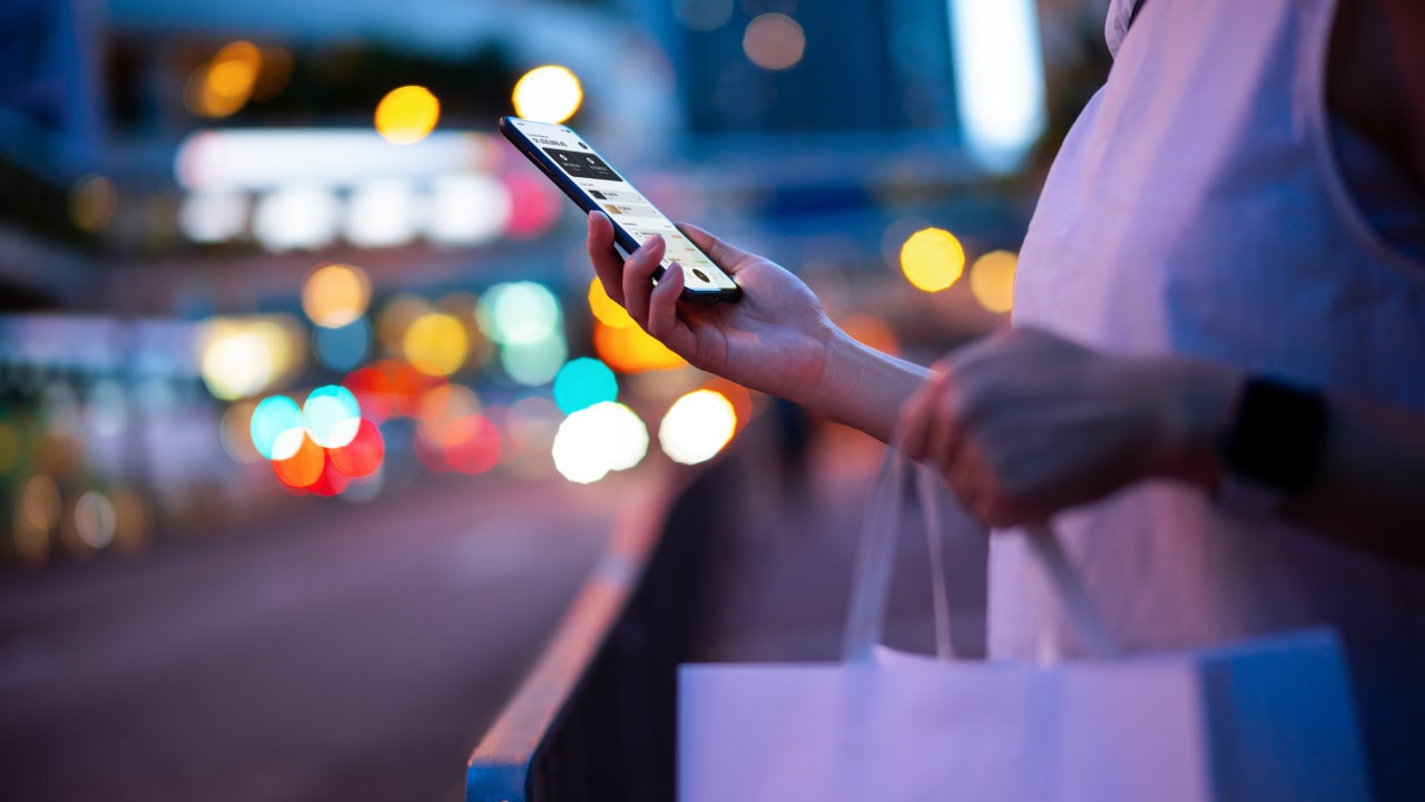 Young woman carrying a paper shopping bag, managing online banking on smartphone in city street against illuminated street lights. Tracking and planning spending. Transferring money, paying bills, checking account balances. Smart banking with technology