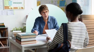 Two women sitting across from each other at a desk. One woman is looking at a piece of paper and the other is explaining something to her.