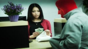 A young, frightened female bank teller places a large wad of cash in a sack as she looks at a bank robber concealed in a red mask, pointing at her in his hoodie pocket.